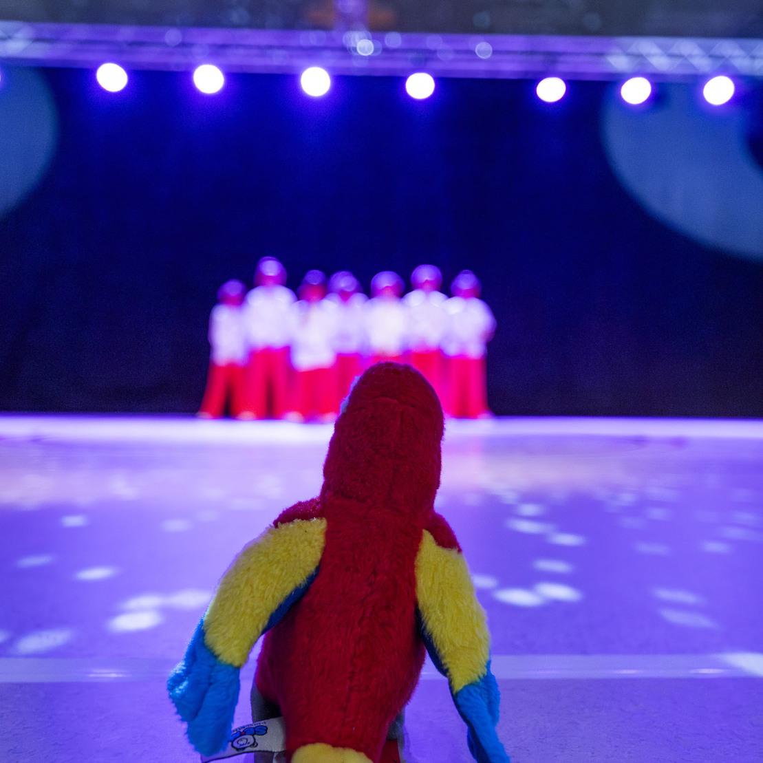 Pl&uuml;schfigur blickt auf eine Tanzgruppe, die auf einer leuchtenden B&uuml;hne mit Discokugel auftritt.
