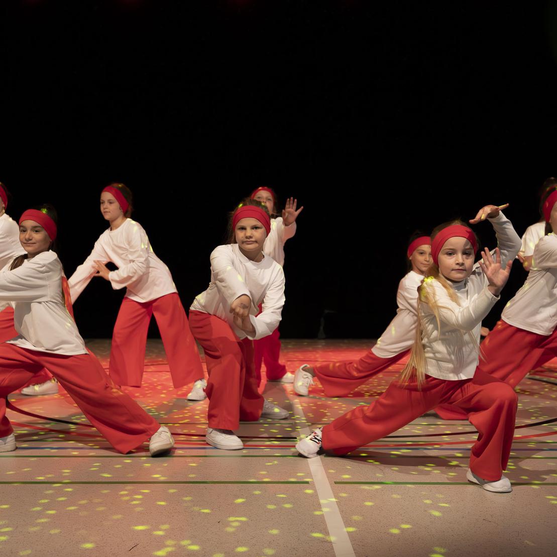 Tanzgruppe von M&auml;dchen in roten Hosen und wei&szlig;en Oberteilen, die synchron auf einer B&uuml;hne performen.