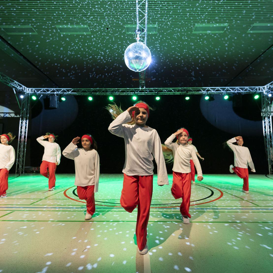 Sechs T&auml;nzer in wei&szlig;en Shirts und roten Hosen f&uuml;hren eine Choreografie unter einer Diskokugel auf einer beleuchteten Fl&auml;che vor.