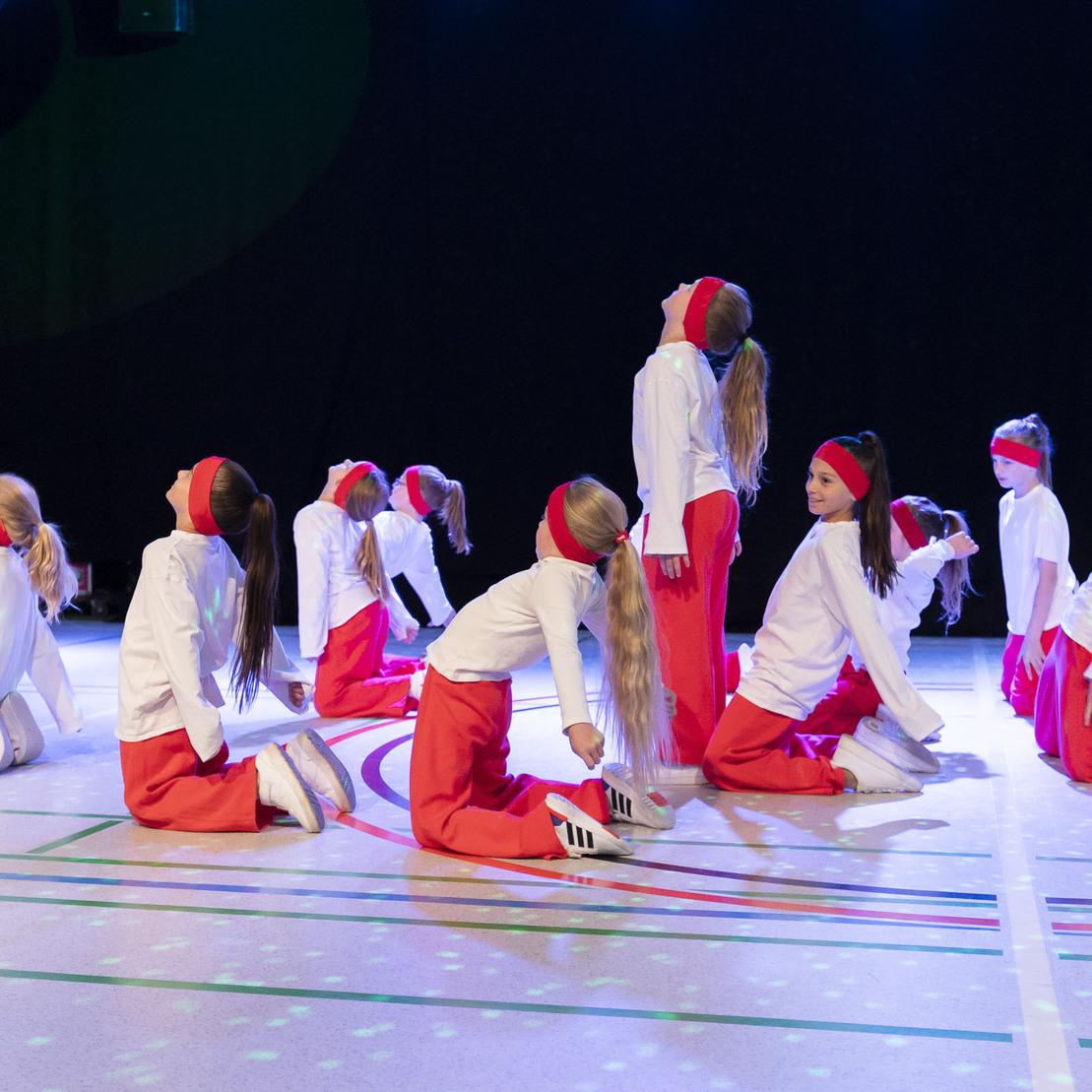 Tanzgruppe in wei&szlig;en Oberteilen und roten Hosen vollf&uuml;hrt eine choreografierte Pose auf einer B&uuml;hnenfl&auml;che.