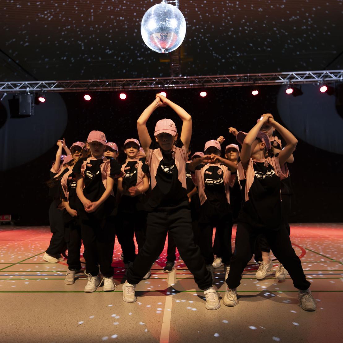 Tanzgruppe in schwarzen Outfits mit pinken Caps, auf einer B&uuml;hne unter Beleuchtung, f&uuml;hrt dynamische Choreografie aus.