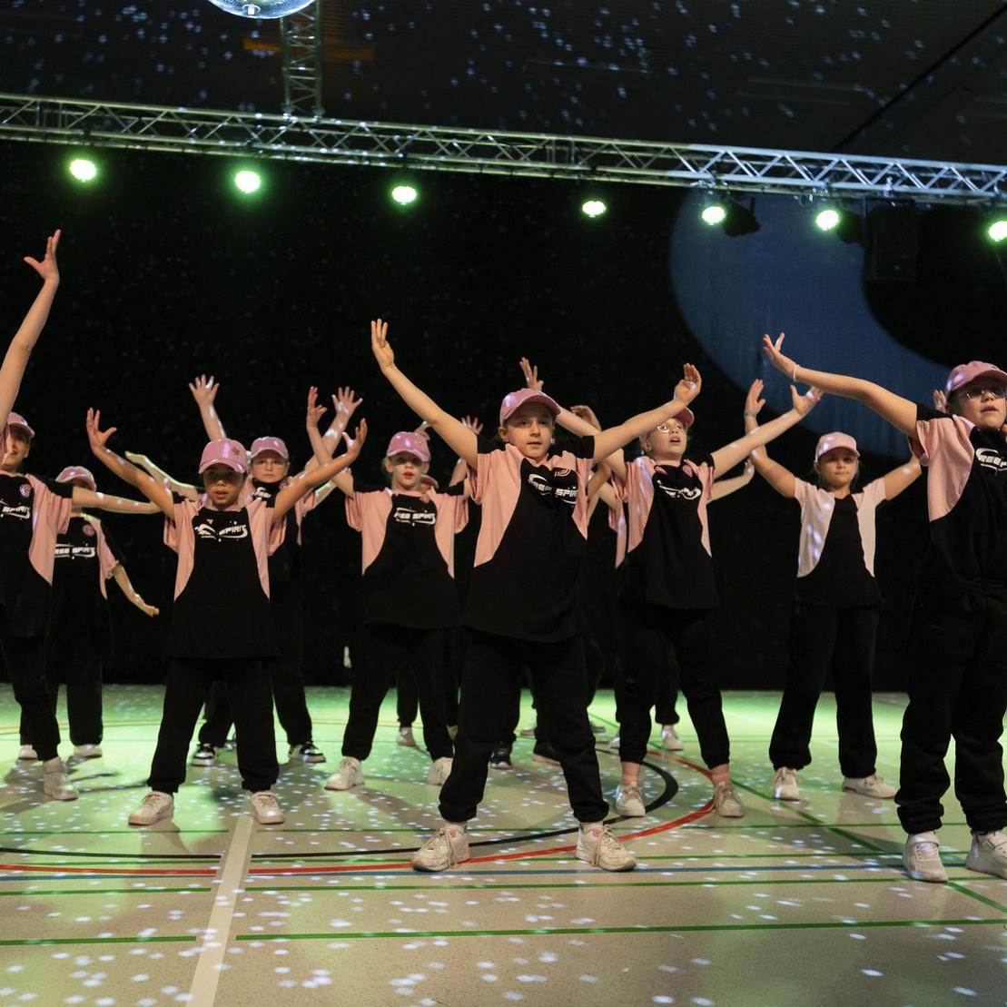 Gruppe junger T&auml;nzer in schwarz-pinker Kleidung f&uuml;hrt eine Choreografie auf einer beleuchteten B&uuml;hne vor.