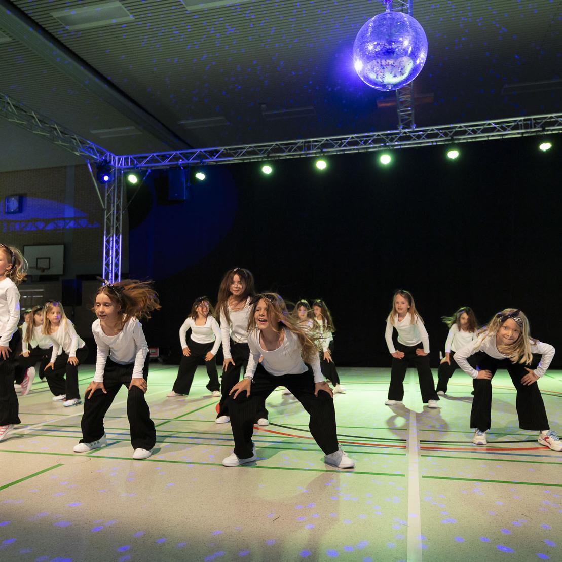 Tanzgruppe von M&auml;dchen in wei&szlig;en Oberteilen und schwarzen Hosen auf einer B&uuml;hne mit Discokugel und Beleuchtung.