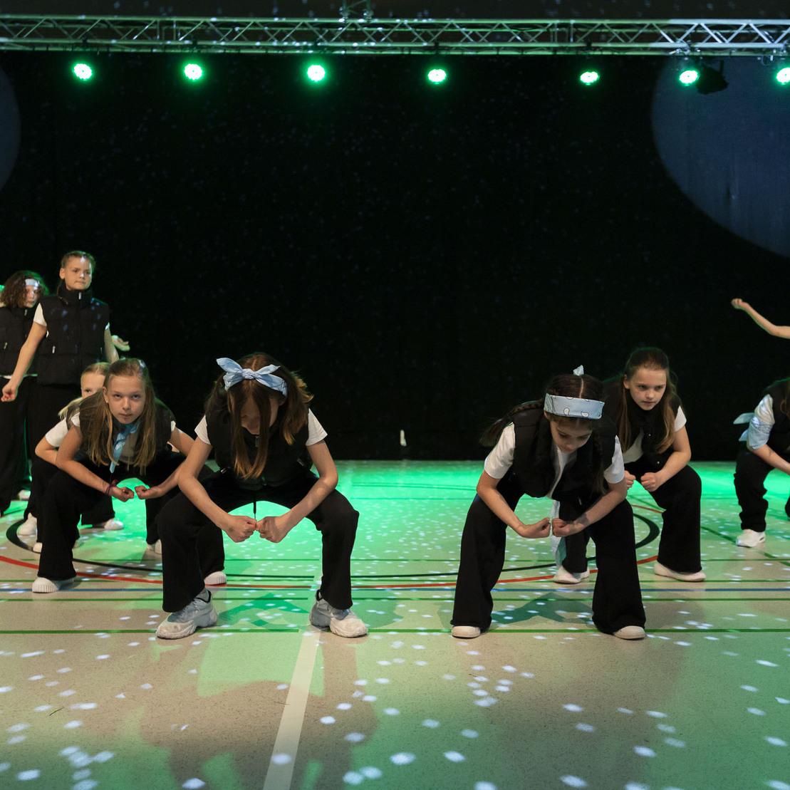 Tanzgruppe in schwarzen Outfits mit blauen Bandanas, die synchron auf einer beleuchteten B&uuml;hne performt.