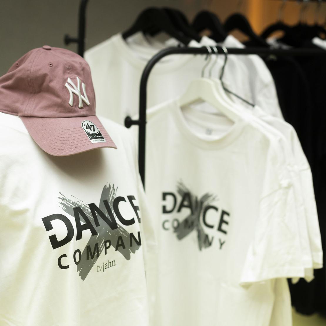 Wei&szlig;es T-Shirt mit "DANCE COMPANY"-Aufdruck und lila Kappe auf einem Kleiderst&auml;nder in einem hellen Raum.