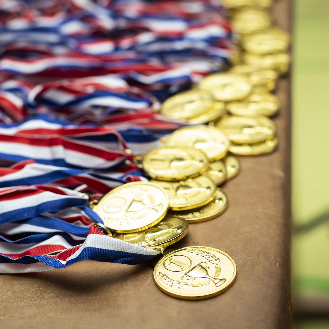 Goldene Medaillen mit rot-blauen B&auml;ndern liegen ordentlich auf einem Tisch in einer Sportumgebung.
