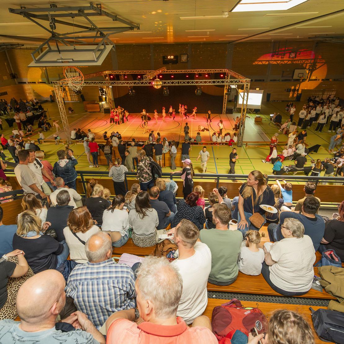 Vogelperspektive auf eine Sporthalle mit Zuschauern, T&auml;nzern auf der B&uuml;hne und festlicher Beleuchtung.