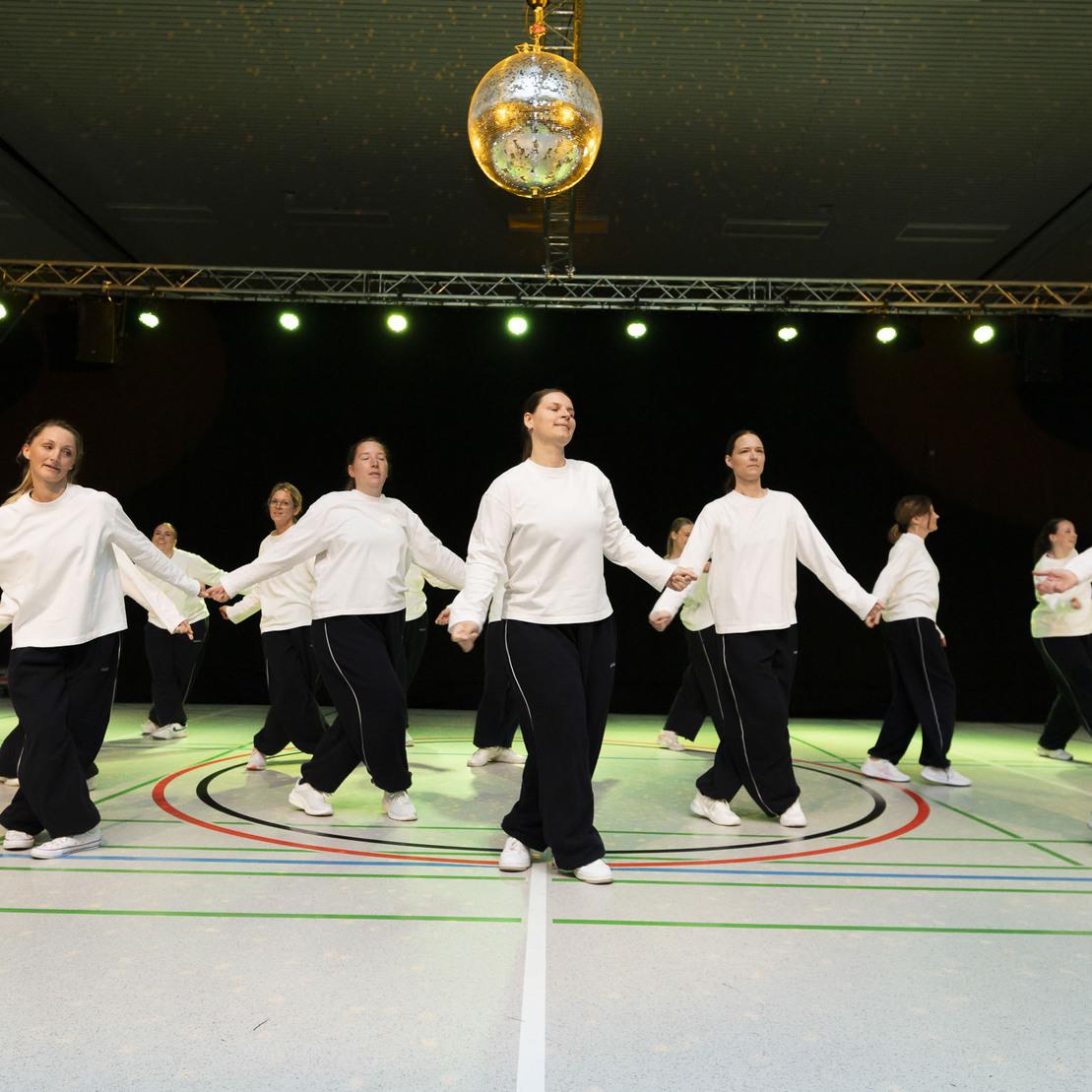 Tanzgruppe in wei&szlig;en Oberteilen und schwarzen Hosen, die synchron auf einer Sportfl&auml;che zu einer Disco-Kugel tanzt.