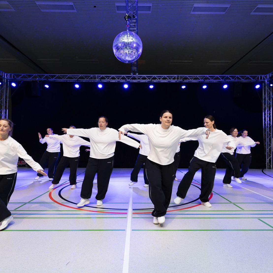 Tanzgruppe in wei&szlig;en Oberteilen und schwarzen Hosen f&uuml;hrt synchron eine Choreografie auf einer beleuchteten B&uuml;hne aus.