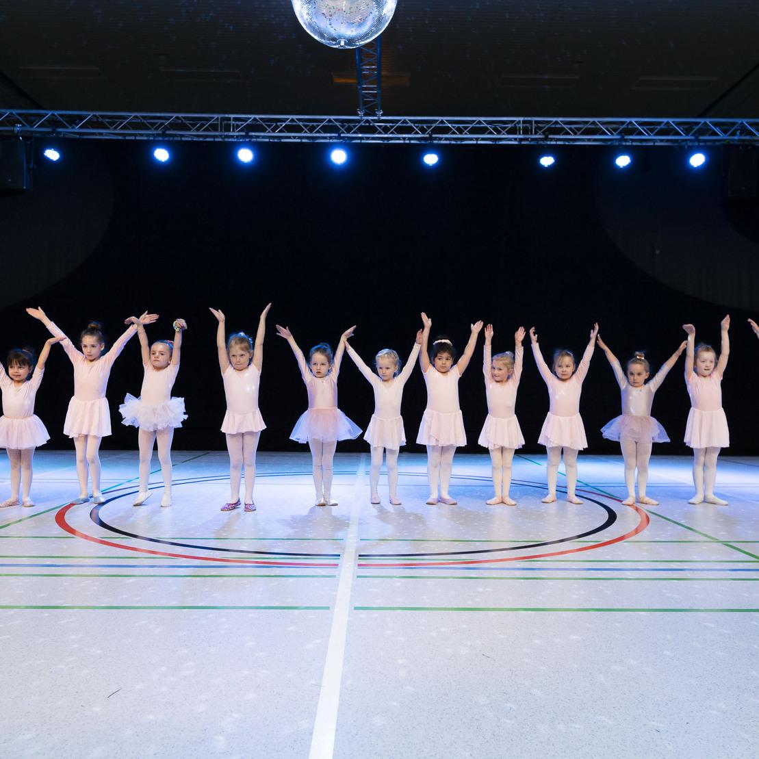 Zehn kleine T&auml;nzerinnen in rosa Ballettkleidern stehen mit erhobenen Armen auf einer beleuchteten B&uuml;hne.