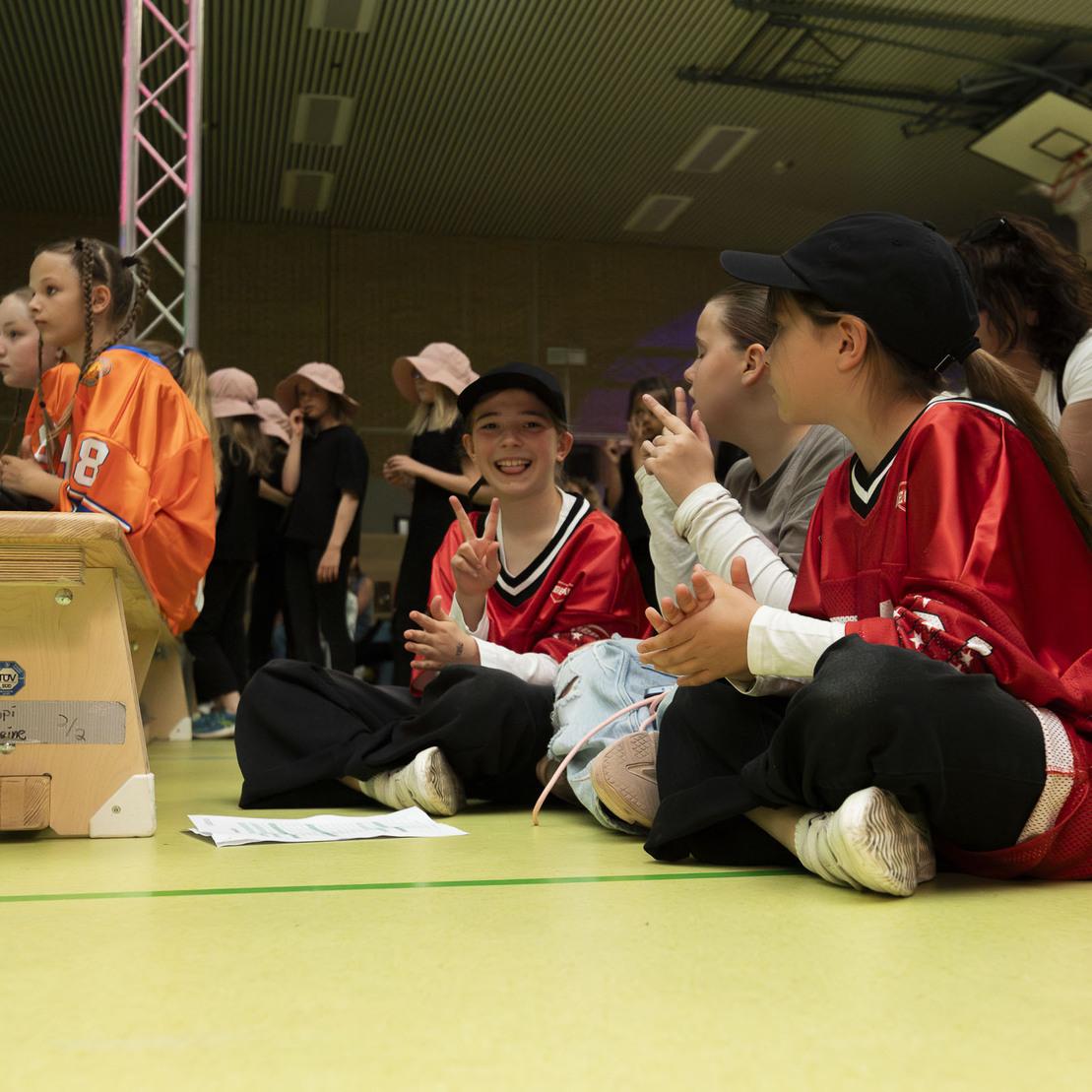 M&auml;dchen in Sportkleidung sitzen l&auml;chelnd auf dem Boden, applaudieren und beobachten eine Veranstaltung in einer Sporthalle.