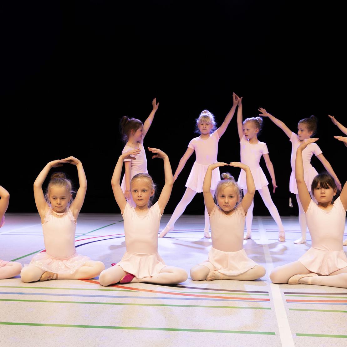 Gruppe von M&auml;dchen in Ballettkleidung, die in verschiedenen Posen auf einer B&uuml;hne stehen und sitzen.