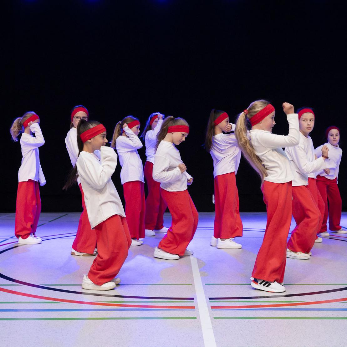Tanzgruppe von M&auml;dchen in roten Hosen und wei&szlig;en Oberteilen f&uuml;hrt eine Choreografie auf einer B&uuml;hne mit buntem Boden aus.