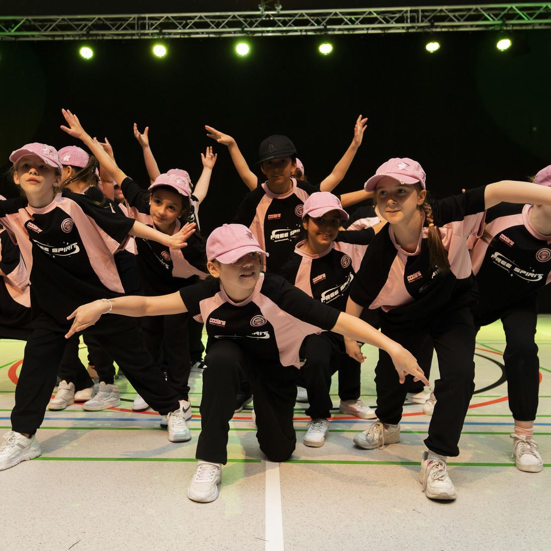 Gruppe von T&auml;nzern in schwarz-pinker Kleidung und M&uuml;tzen, die synchron auf einer B&uuml;hne posiert.