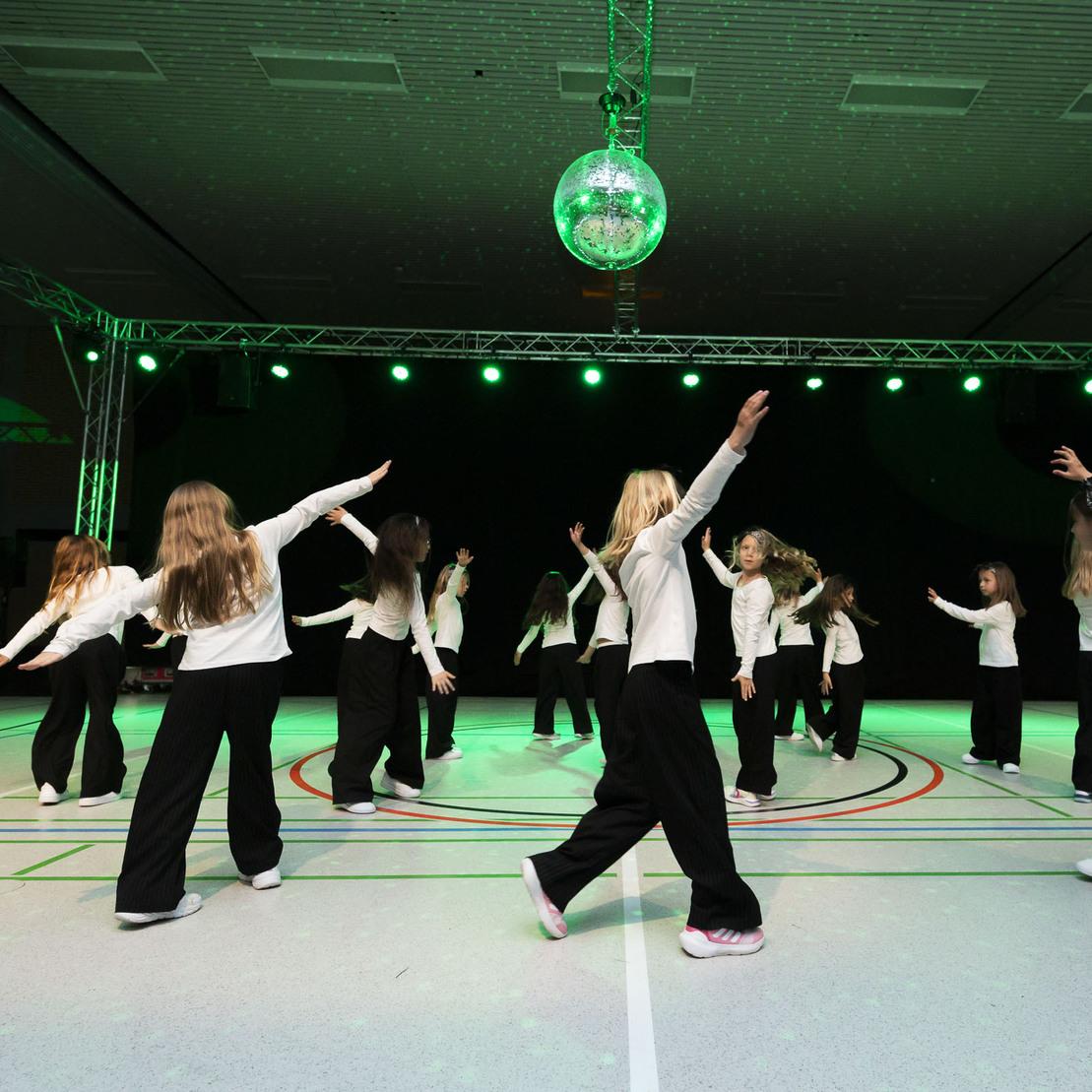 Tanzende Kinder in schwarz-wei&szlig;er Kleidung auf einer beleuchteten Sporthalle mit einem Discokugel &uuml;ber der B&uuml;hne.