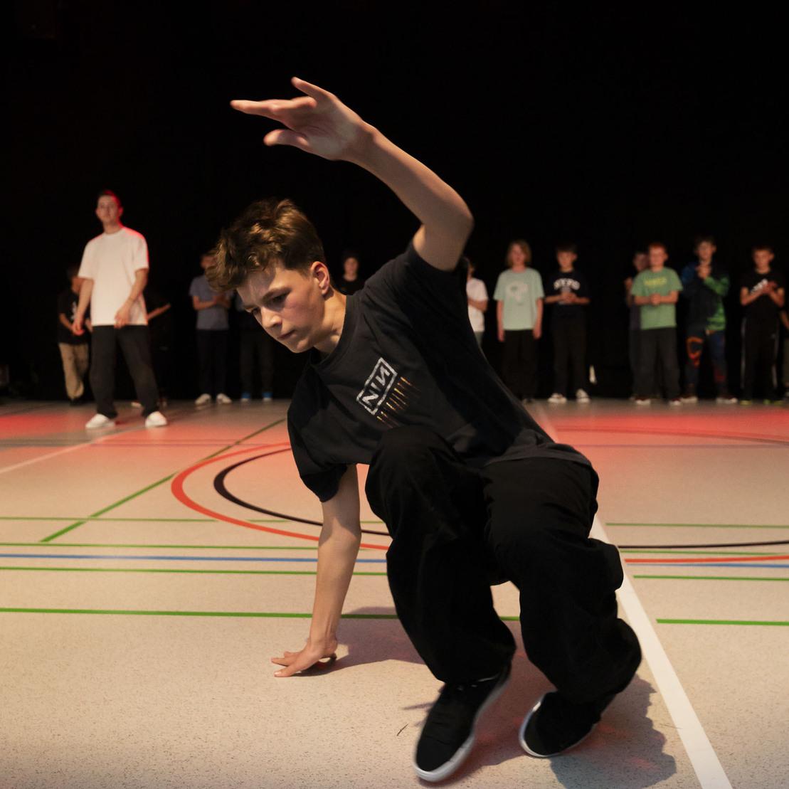 Junger T&auml;nzer in der Aktion beim Breakdance auf einem Sportboden, Zuschauer stehen im Hintergrund.