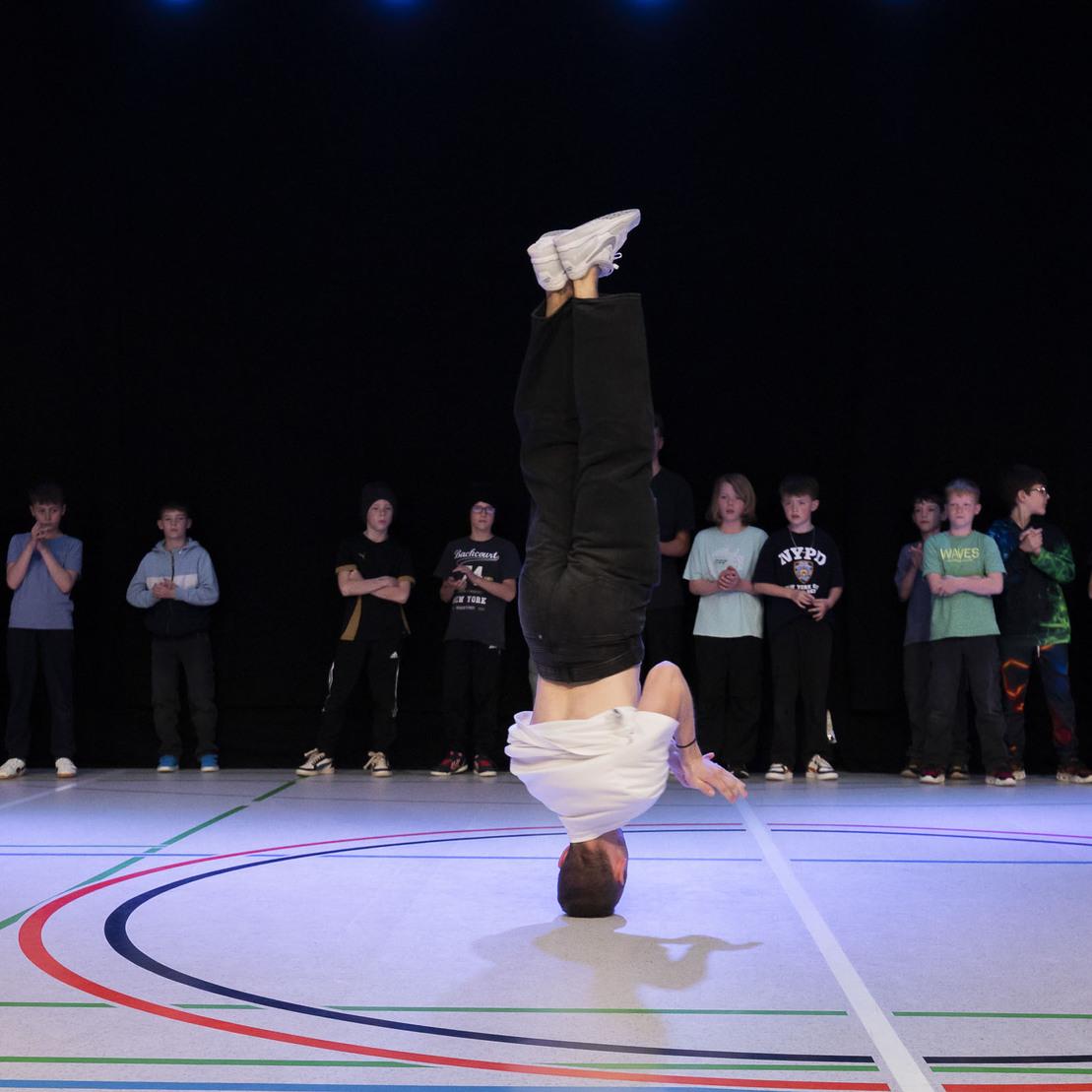 Ein T&auml;nzer f&uuml;hrt einen Handstand auf einer Sportfl&auml;che aus, w&auml;hrend eine Gruppe von Jugendlichen zuschaut.