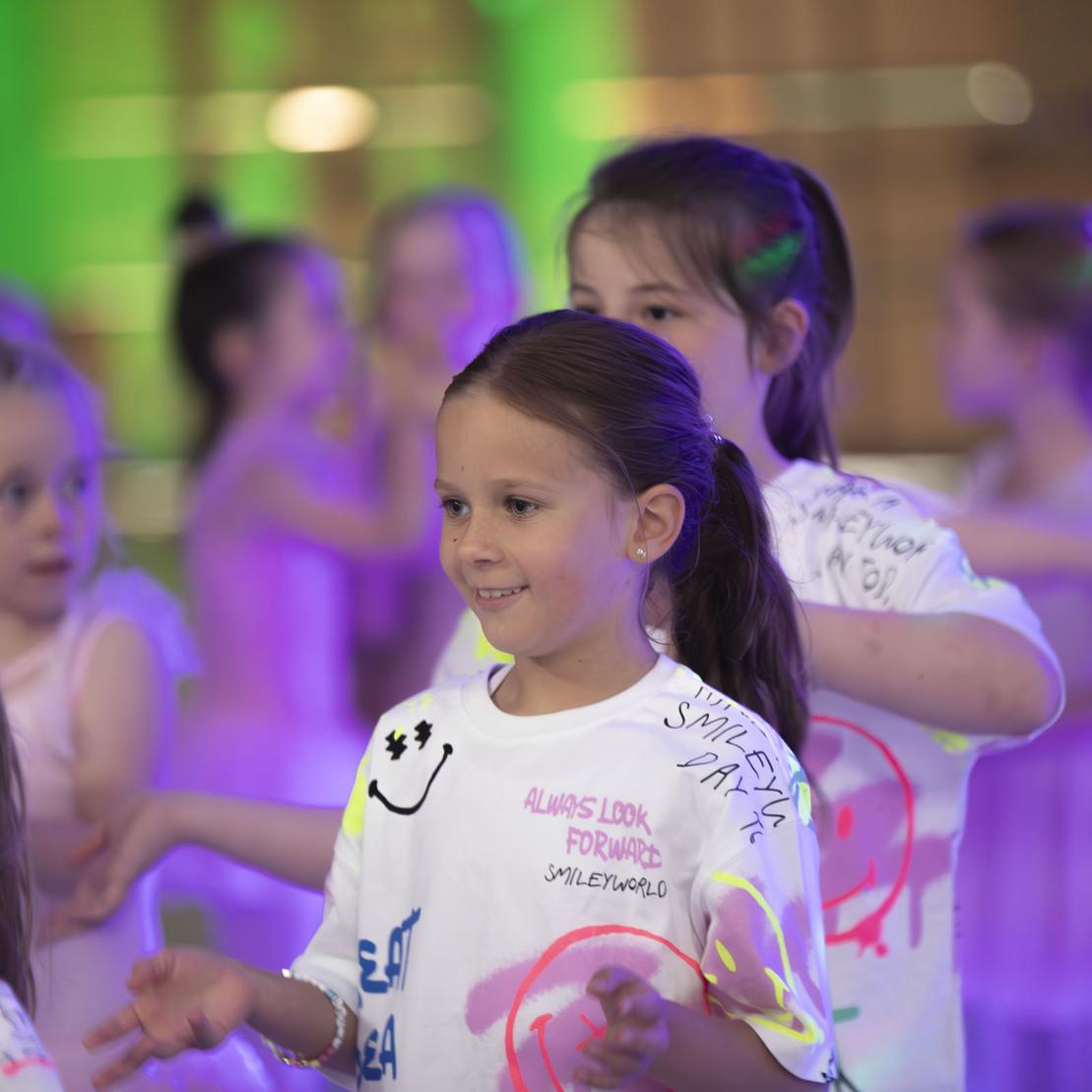 M&auml;dchen mit einem L&auml;cheln tanzt in einem bunten T-Shirt zusammen mit anderen Kindern in einem beleuchteten Raum.