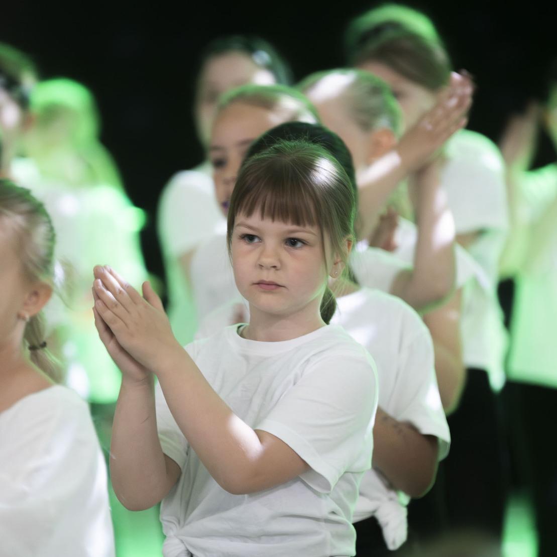 M&auml;dchen mit braunen Haaren klatscht in den H&auml;nden, umgeben von anderen Kindern in wei&szlig;en T-Shirts bei einer Auff&uuml;hrung.