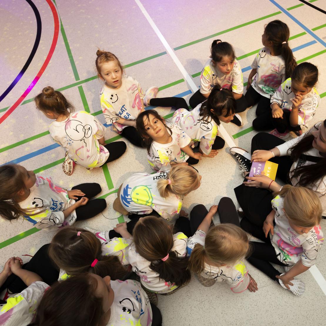 Gruppe von Kindern in bunten T-Shirts sitzt auf dem Boden eines Sportplatzes und h&ouml;rt einer erwachsenen Person zu.
