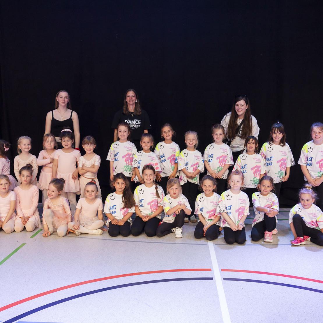 Gruppe von Kindern in Tanzkleidung und T-Shirts, posierend mit Betreuern auf einer Sportfl&auml;che vor schwarzem Hintergrund.