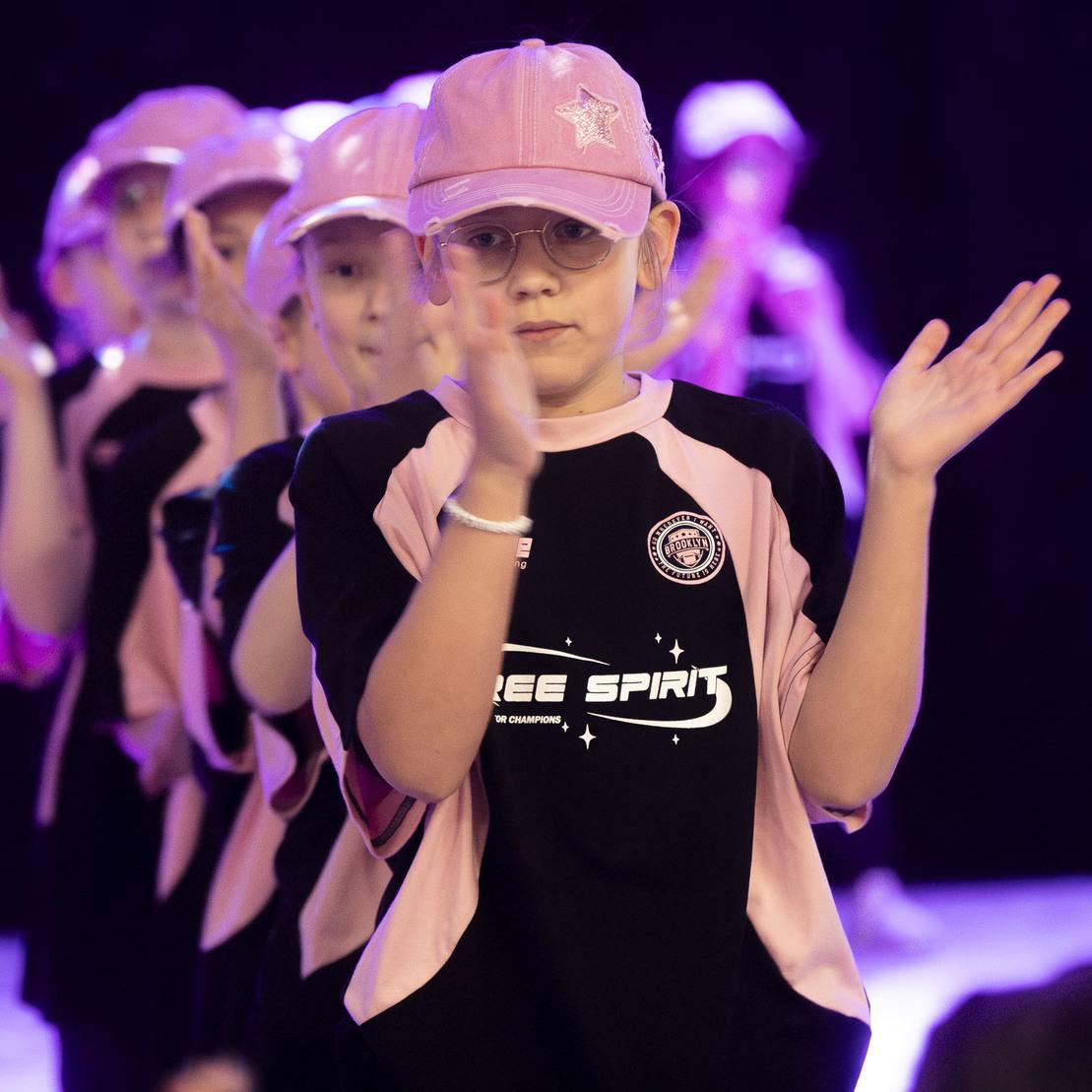 Gruppen von Kinder in rosa Kappen und T-Shirts klatschen w&auml;hrend einer Tanzauff&uuml;hrung auf einer beleuchteten B&uuml;hne.
