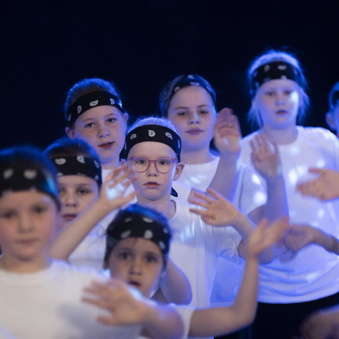 Gruppe von Kindern in wei&szlig;en T-Shirts und schwarzen Bandanas bei einer Tanzauff&uuml;hrung mit beleuchtetem Hintergrund.