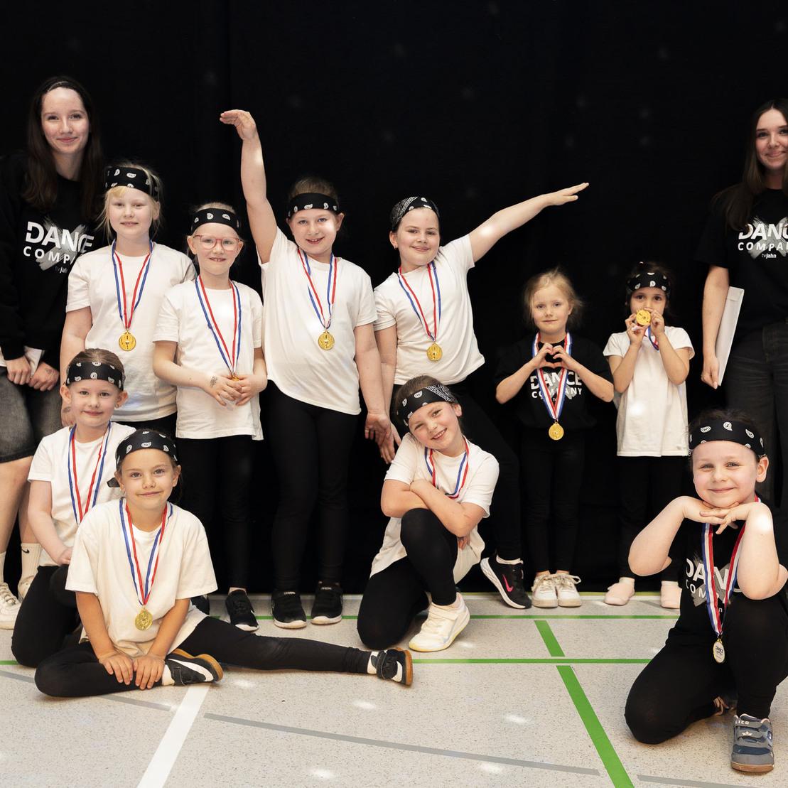 Gruppe von tanzenden Kindern in T-Shirts mit Medaillen, posierend mit zwei Trainerinnen auf einer B&uuml;hne.