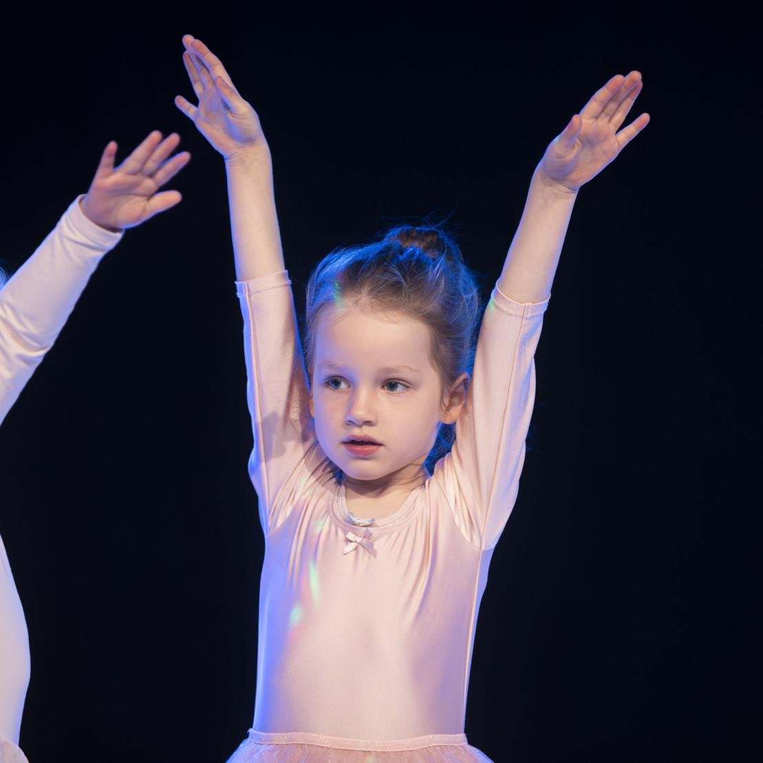 M&auml;dchen in Ballettkleidung mit erhobenen Armen steht auf einer B&uuml;hne, umgeben von weiteren T&auml;nzerinnen.