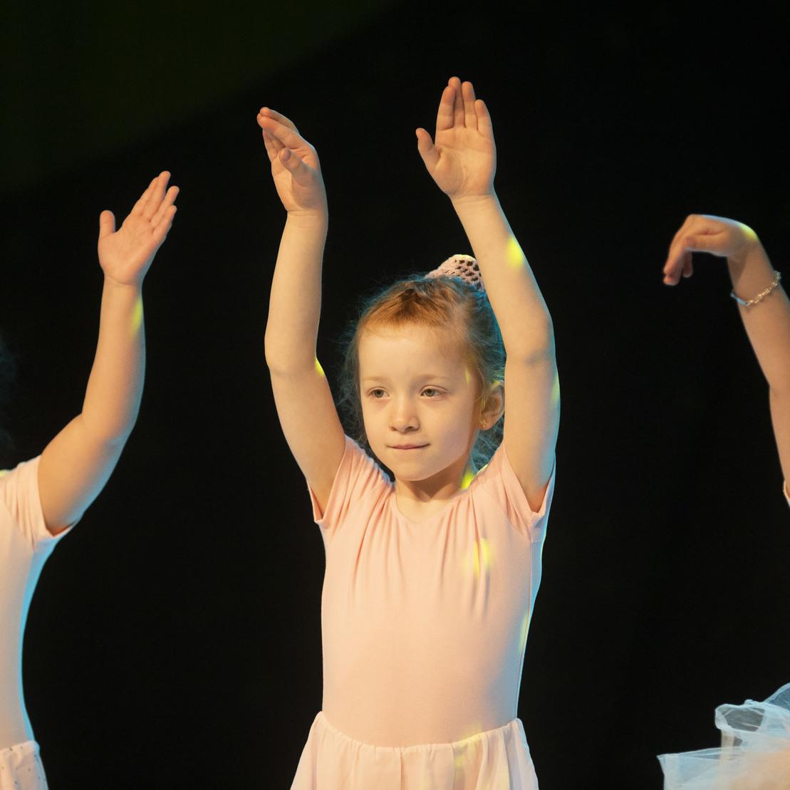 Drei kleine Ballettt&auml;nzerinnen in rosa Kost&uuml;men heben die Arme vor einem dunklen Hintergrund.