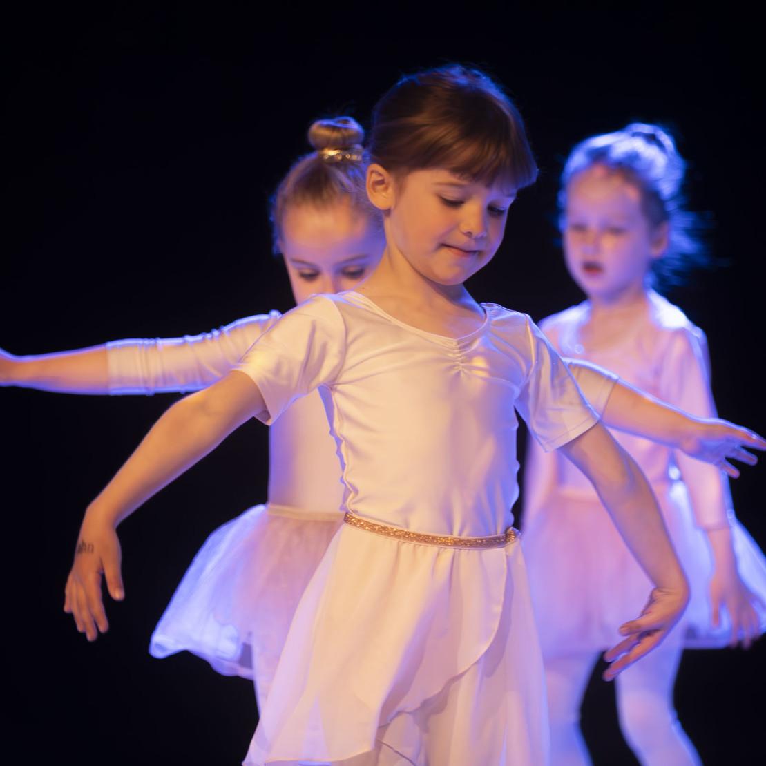 Drei kleine M&auml;dchen in Ballettkleidern tanzen auf einer dunklen B&uuml;hne mit sanfter Beleuchtung.