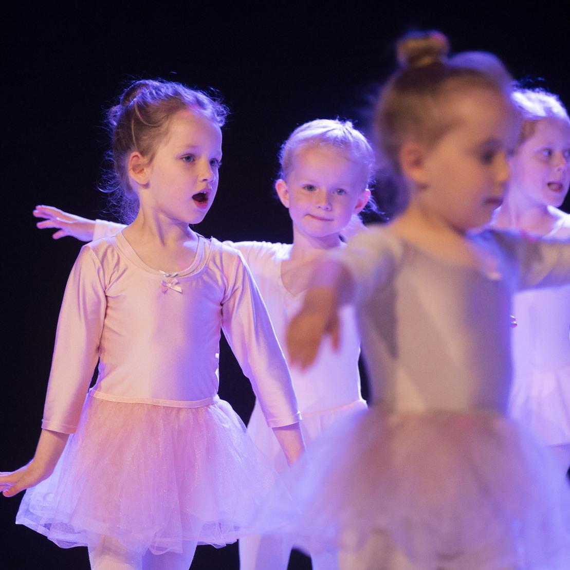 Vier kleine Ballettt&auml;nzerinnen in rosa T&uuml;llr&ouml;cken f&uuml;hren gemeinsam eine Choreografie auf einer dunklen B&uuml;hne vor.