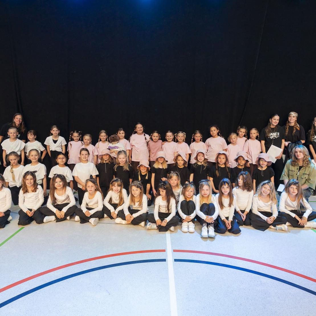 Gruppennaufnahme von tanzenden Kindern in Sportkleidung auf einem Hallenboden mit bunten Linien.