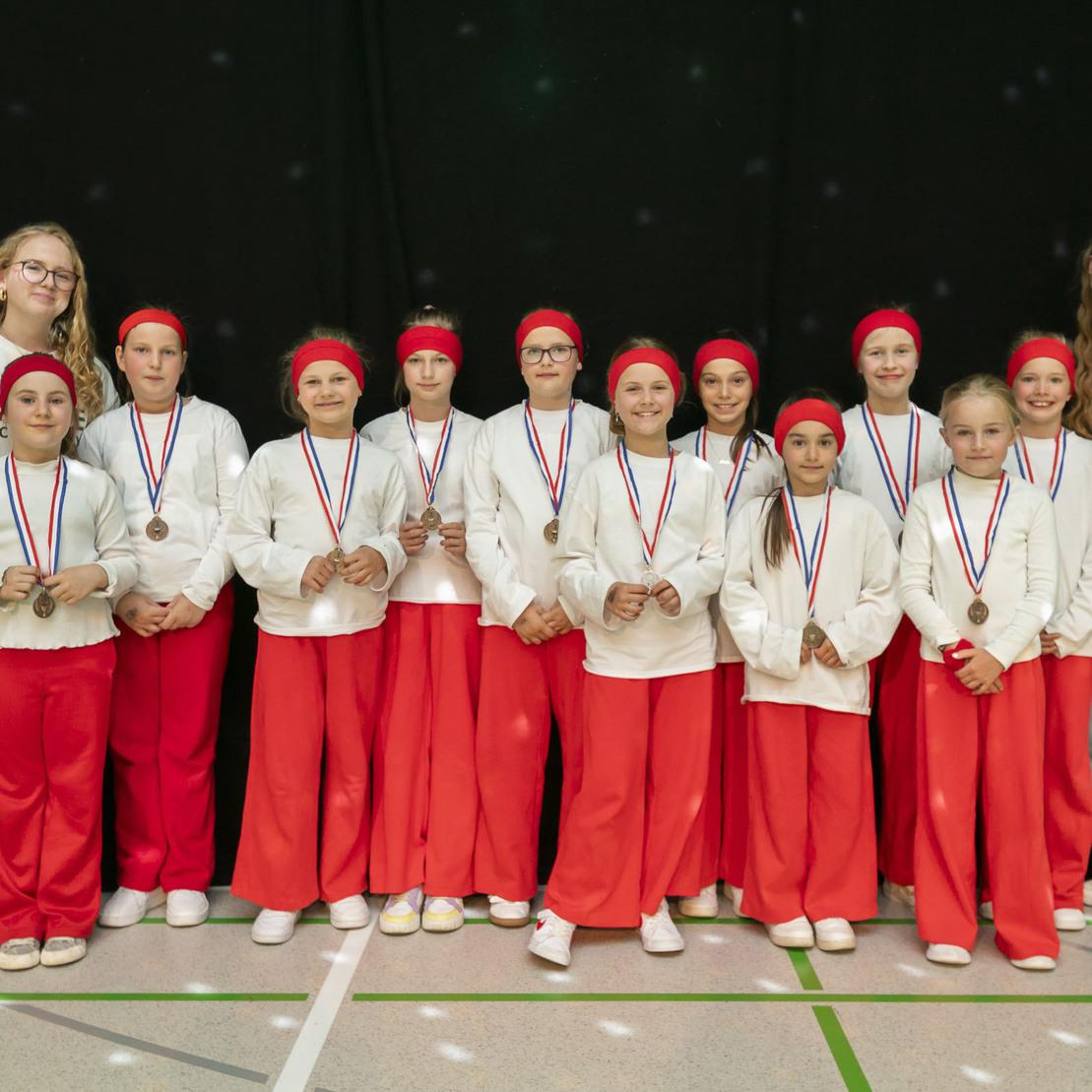 Gruppenfoto von zw&ouml;lf Kindern in roten Hosen und wei&szlig;en Oberteilen mit Medaillen, umrahmt von zwei Betreuern.