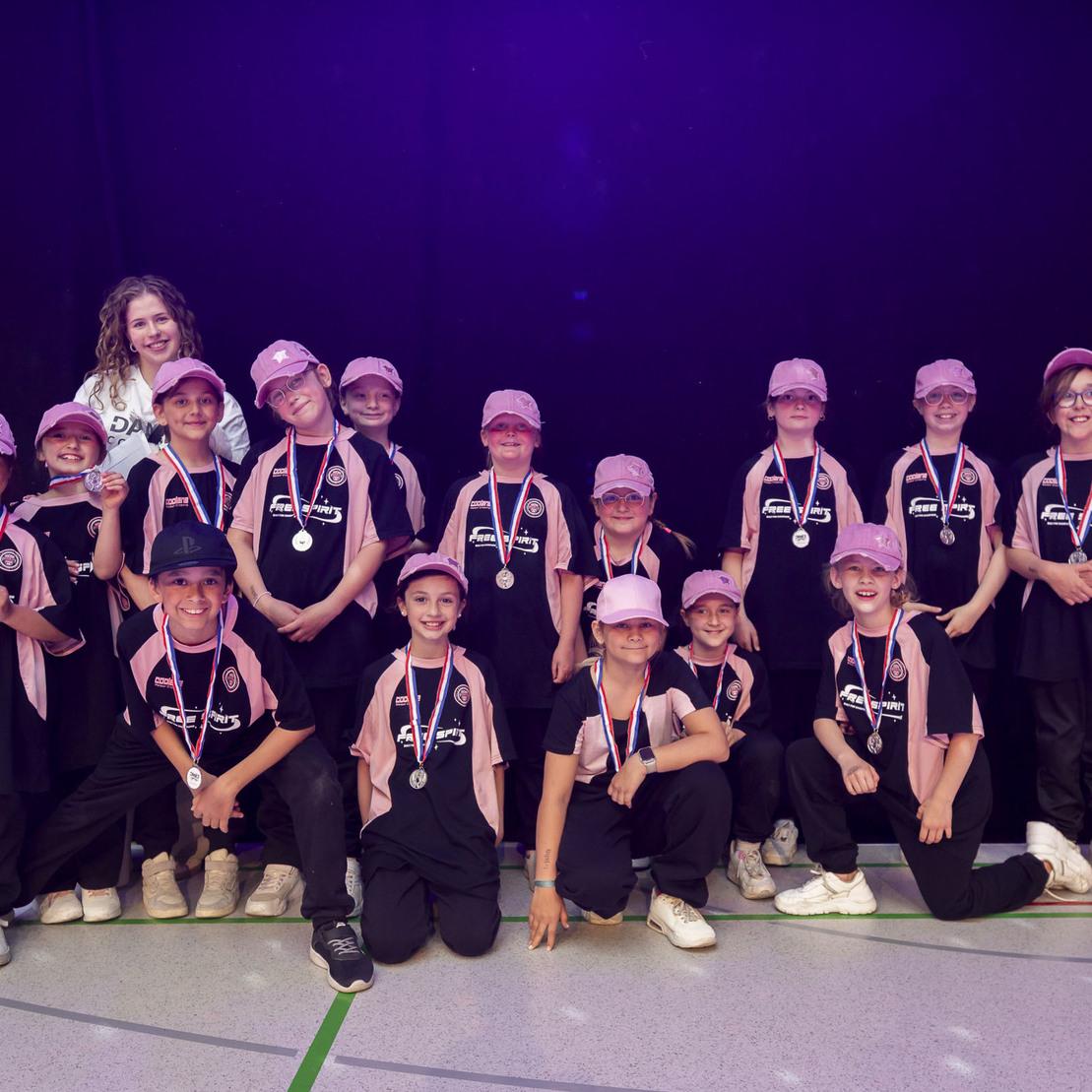 Gruppe von 16 Kindern in Sportkleidung mit Medaillen und pinken Kappen, posierend vor einem dunklen Hintergrund.