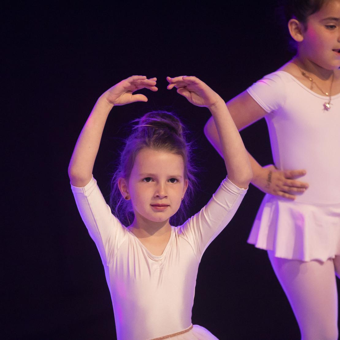 Zwei M&auml;dchen in Ballettkleidung tanzen auf einer dunklen B&uuml;hne, eines formt eine Tanzpose mit erhobenen Armen.