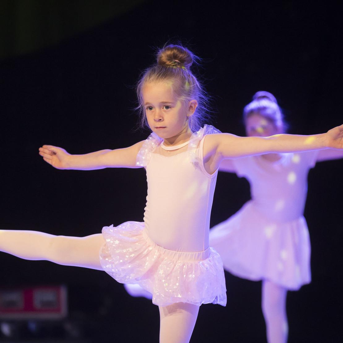 M&auml;dchen in einem wei&szlig;en Ballettkost&uuml;m tanzt auf einer B&uuml;hne, ein Bein seitlich gestreckt, mit anderen T&auml;nzerinnen im Hintergrund.