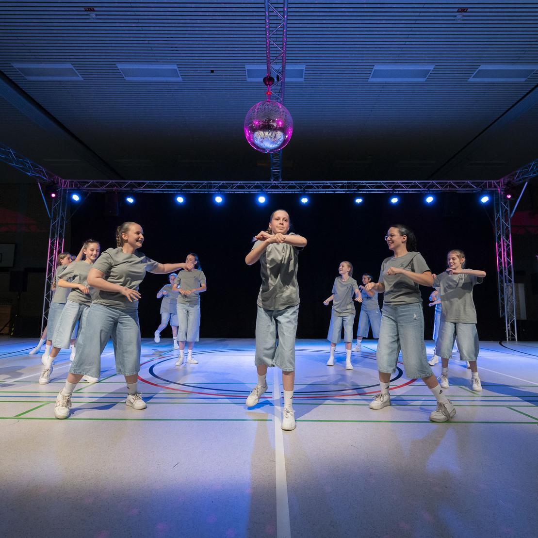Tanzgruppe in grauen Outfits f&uuml;hrt eine Choreografie unter bunten Lichtern und einer Discokugel auf einer Sportfl&auml;che vor.