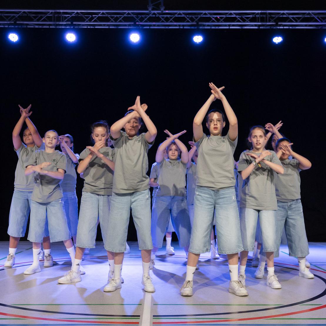 Gruppe von T&auml;nzern in grauen Shirts und Jean-Shorts, die synchron auf einer B&uuml;hne mit B&uuml;hnenlichtern tanzen.