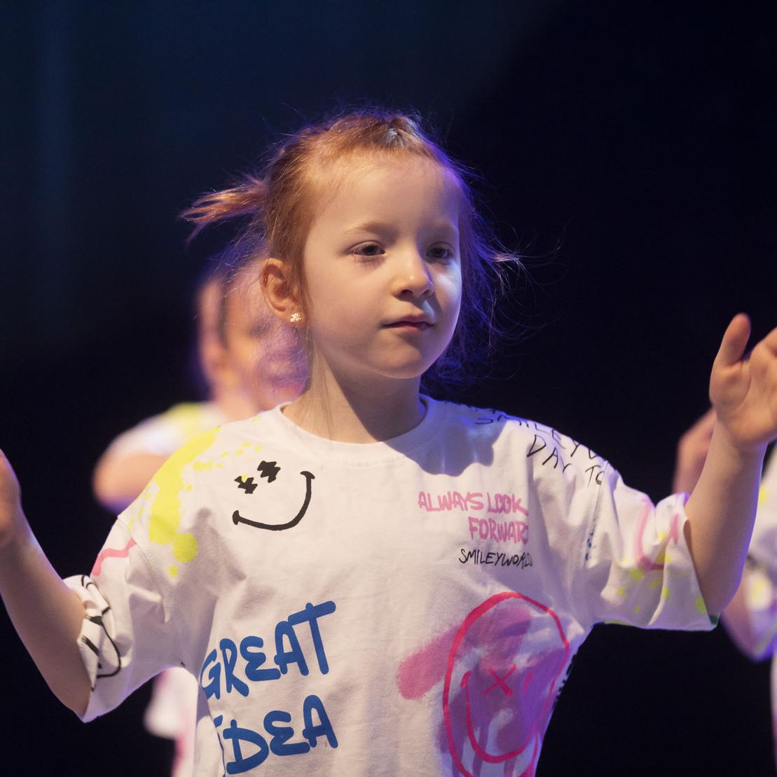 M&auml;dchen in buntem T-Shirt tanzt auf einer B&uuml;hne, umgeben von anderen Kindern mit &auml;hnlicher Kleidung.