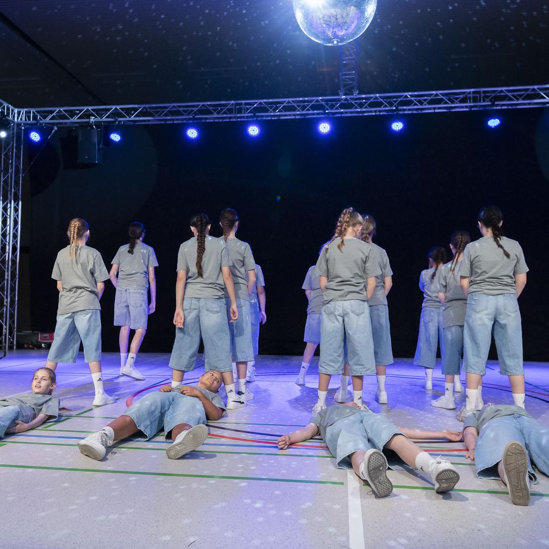 Tanzgruppe in grauen Outfits auf B&uuml;hne, einige T&auml;nzer liegen auf dem Boden, w&auml;hrend andere stehen. Diskokugel im Hintergrund.