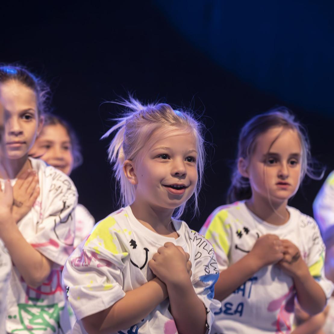 Gruppe von Kindern in bunten T-Shirts, die mit Freude eine Tanzauff&uuml;hrung auf der B&uuml;hne pr&auml;sentieren.