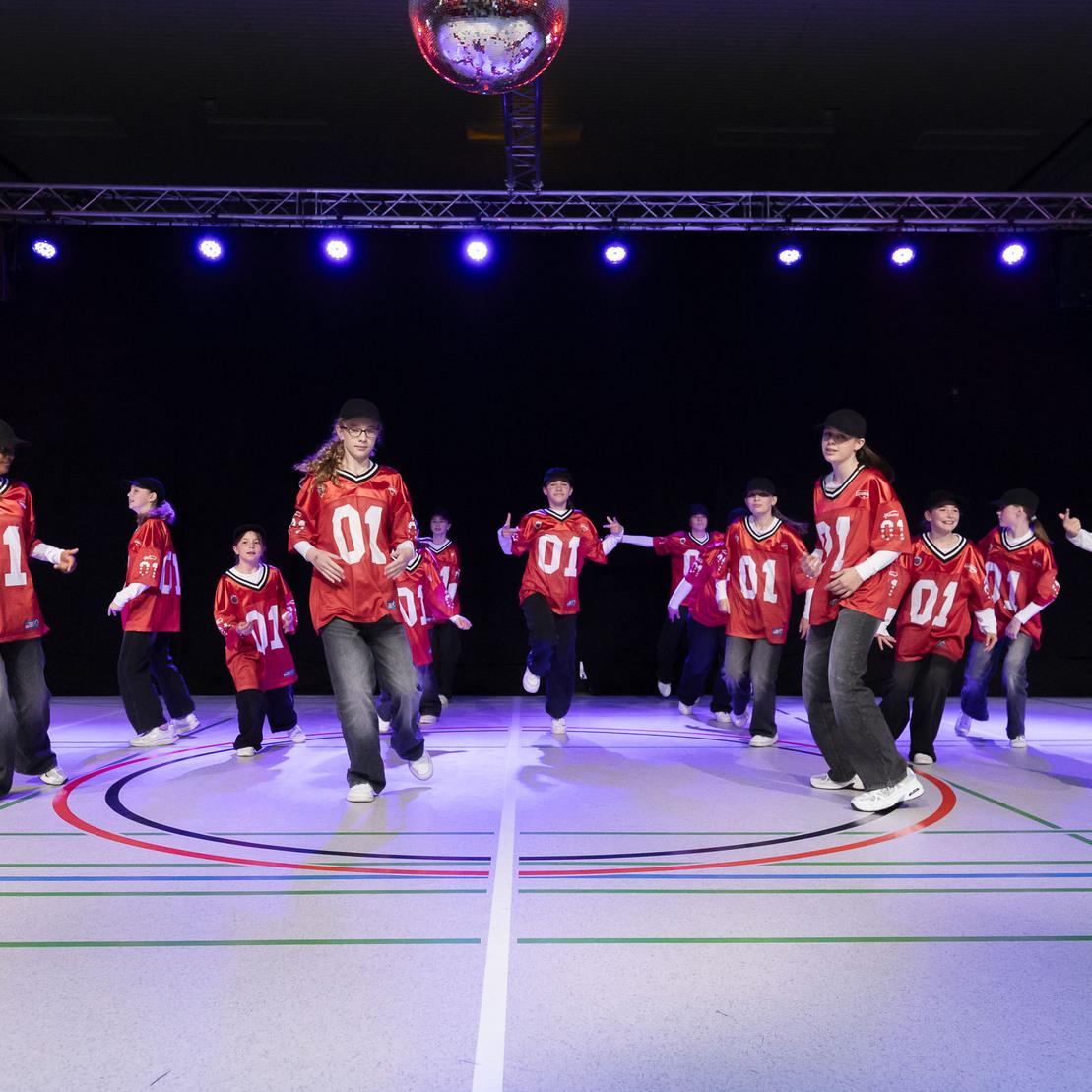 Tanzgruppe mit roten Shirts auf einer bunten Sportfl&auml;che, beleuchtet von farbigem Licht w&auml;hrend einer Auff&uuml;hrung.