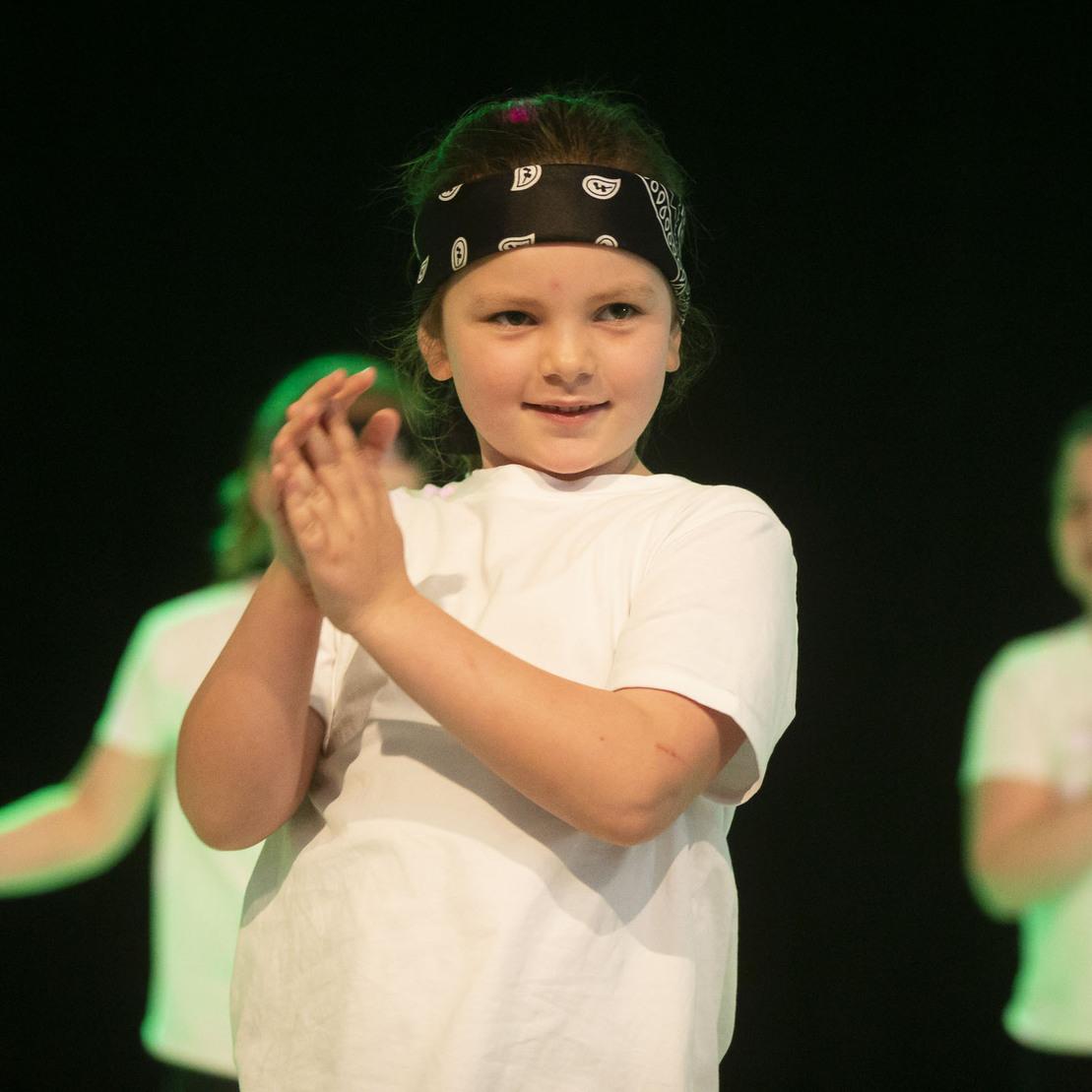Kind mit Bandana und wei&szlig;em T-Shirt f&uuml;hrt einen Tanz auf einer B&uuml;hne vor, im Hintergrund zwei weitere Kinder clappen.