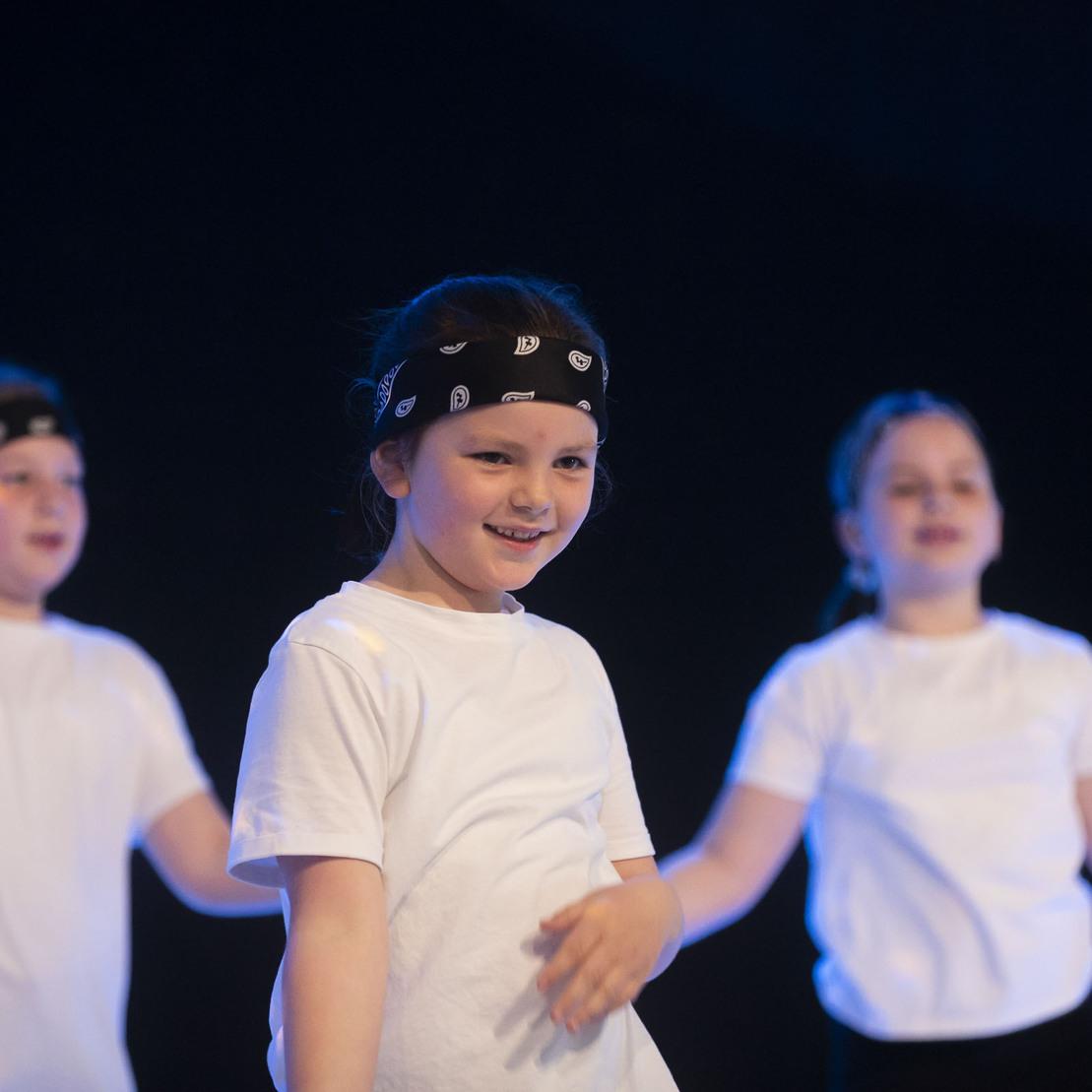 Drei Kinder in wei&szlig;en Shirts und Bandanas tanzen auf einer B&uuml;hne mit blauer Beleuchtung.