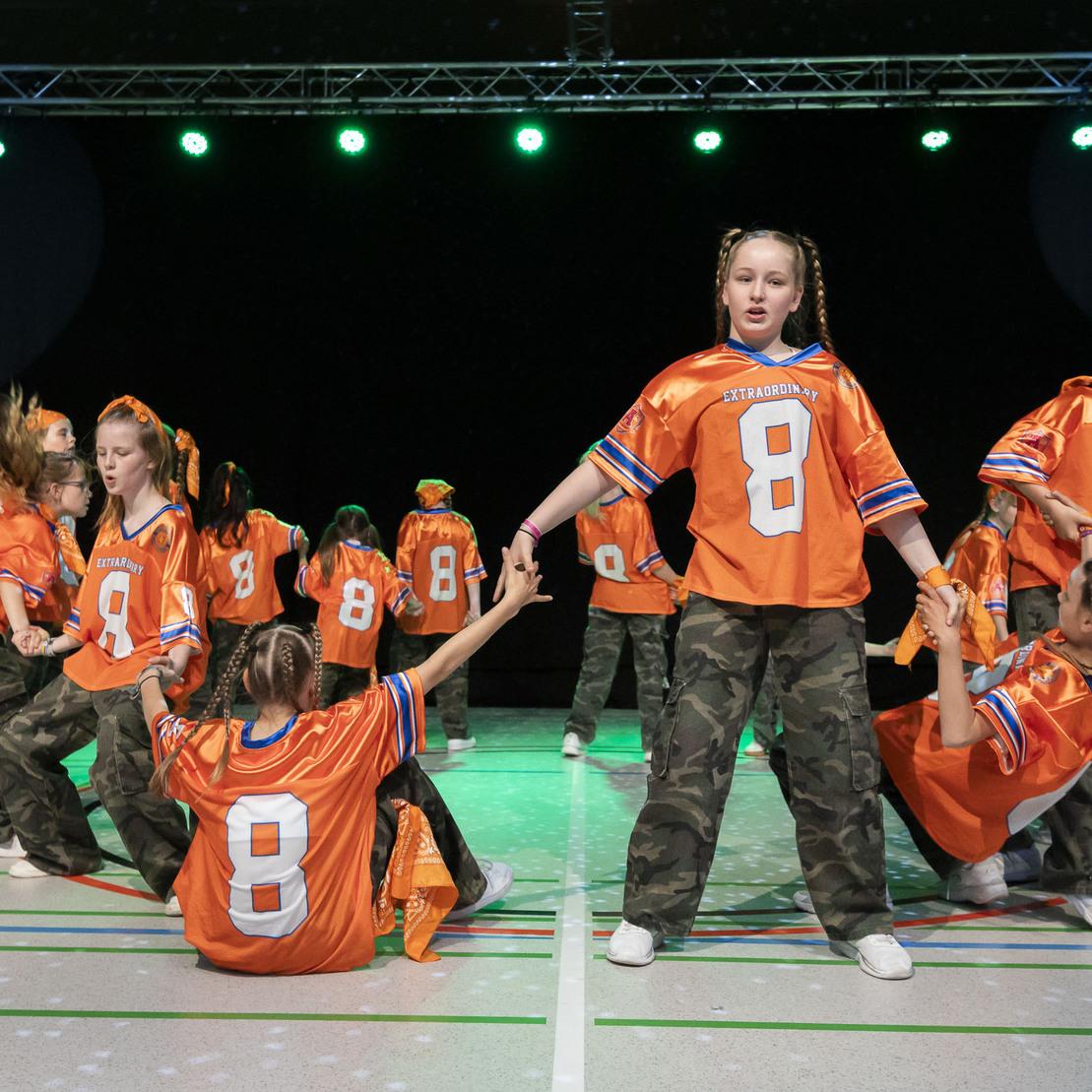 Tanzgruppe in orangefarbenen Trikots mit der Zahl 8, die auf einer B&uuml;hne mit gr&uuml;nem Licht auftritt.