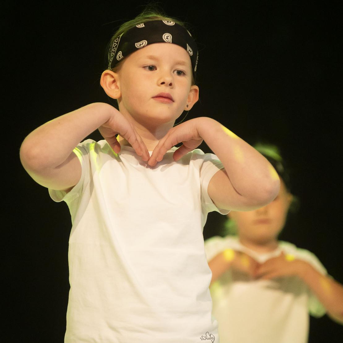 Kinder tanzen auf einer B&uuml;hne, eines tr&auml;gt ein schwarzes Bandana und hat die H&auml;nde in Brusth&ouml;he platziert.