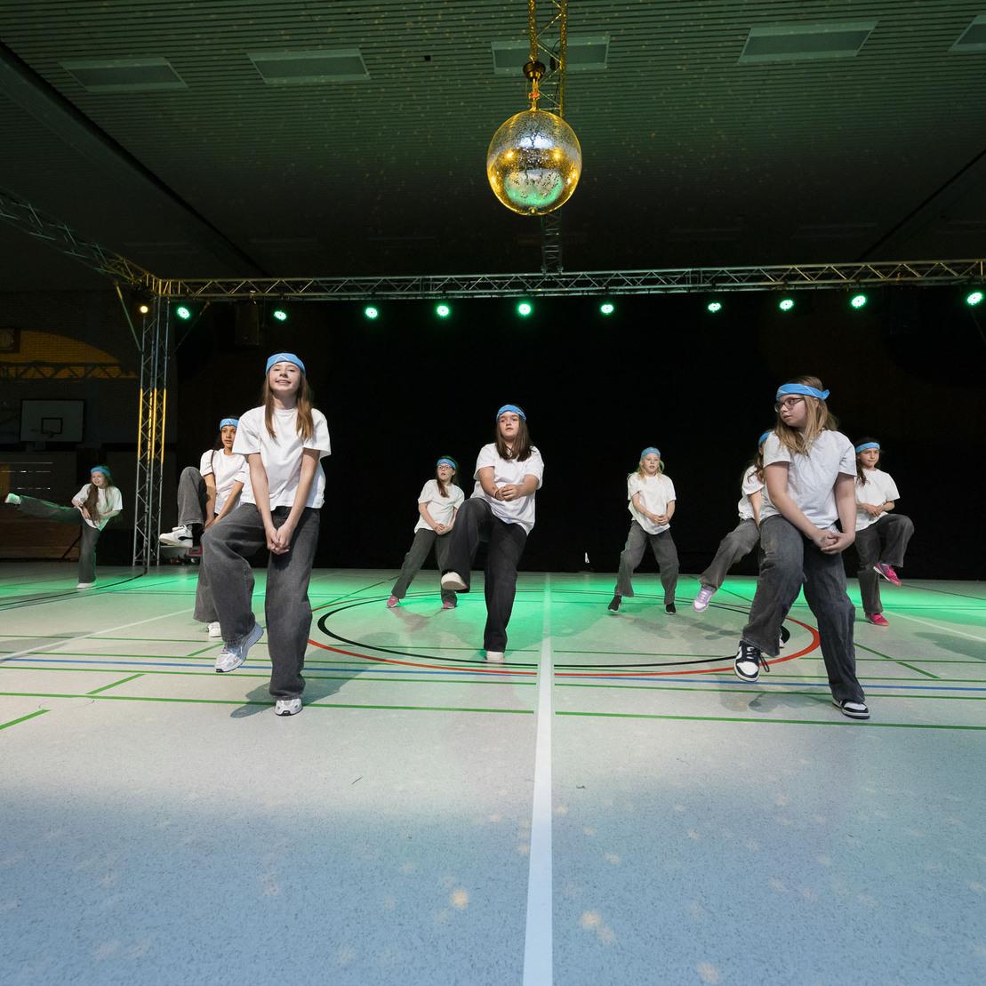 Tanzgruppe in sportlicher Kleidung f&uuml;hrt eine Choreografie unter einem Discokugel-Licht in einer Halle aus.