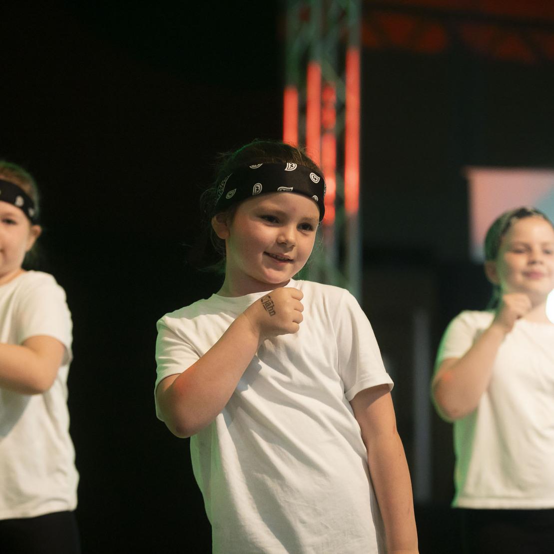 Drei Kinder in wei&szlig;en T-Shirts und Stirnb&auml;ndern f&uuml;hren eine Choreografie auf einer B&uuml;hne vor.