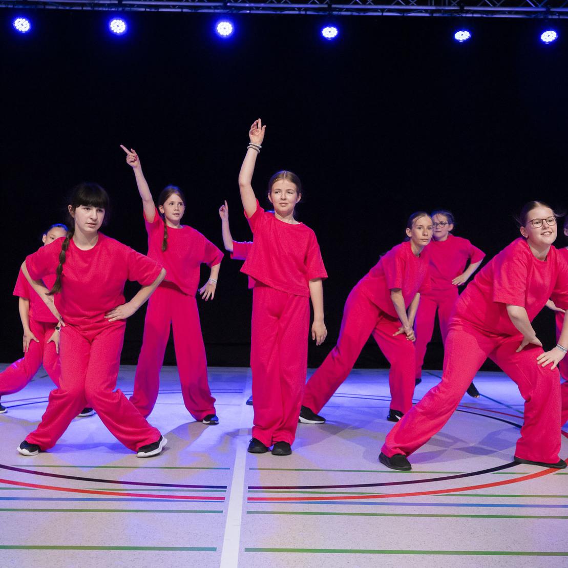 Gruppentanz von sieben M&auml;dchen in roten Outfits auf einer Sportfl&auml;che mit bunten Linien und blauem Scheinwerferlicht.